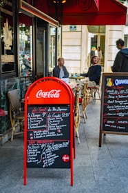 Outdoor seating area of a café with two men seated at a table, and another person standing nearby. The scene includes a red Coca-Cola branded menu board displaying breakfast options in both French and English. A secondary menu board is present on the right, listing various dishes. The café has a classic Parisian look with wicker chairs and a red awning.