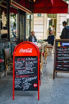 Outdoor seating area of a café with two men seated at a table, and another person standing nearby. The scene includes a red Coca-Cola branded menu board displaying breakfast options in both French and English. A secondary menu board is present on the right, listing various dishes. The café has a classic Parisian look with wicker chairs and a red awning.