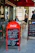 Outdoor seating area of a café with two men seated at a table, and another person standing nearby. The scene includes a red Coca-Cola branded menu board displaying breakfast options in both French and English. A secondary menu board is present on the right, listing various dishes. The café has a classic Parisian look with wicker chairs and a red awning.