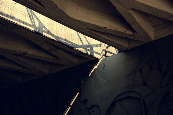 Concrete beams form an intricate architectural structure with strong shadows cast by beams and light filtering through. Patterns and shapes in the concrete create a dynamic visual composition.