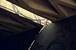 Concrete beams form an intricate architectural structure with strong shadows cast by beams and light filtering through. Patterns and shapes in the concrete create a dynamic visual composition.