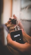 A person holds a vaping device emitting smoke. The device has a digital display with various settings. The hand grips the device firmly, emphasizing the act of vaping.