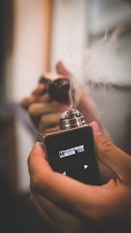 A person holds a vaping device emitting smoke. The device has a digital display with various settings. The hand grips the device firmly, emphasizing the act of vaping.
