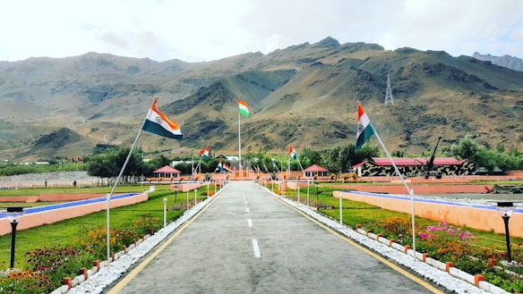 A landscaped area with a paved walkway lined with flowers and grass, bordered by Indian flags on poles. In the background, there are mountains and some structures with camouflaged patterns. The sky is partly cloudy, and the scene conveys a sense of national pride.