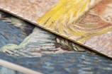 Close-up view of two textured, overlapping canvases displaying vibrant brushstrokes. One canvas features swirling patterns of yellow and beige, while the other showcases a mix of blue and orange tones. The detailed texture of the canvas is visible, highlighting the artistic style.