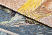 Close-up view of two textured, overlapping canvases displaying vibrant brushstrokes. One canvas features swirling patterns of yellow and beige, while the other showcases a mix of blue and orange tones. The detailed texture of the canvas is visible, highlighting the artistic style.