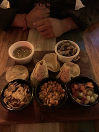 A wooden table holds a platter with a variety of snacks, including nuts, crackers, crispy snacks, and a small bowl of chutney. A pair of hands, likely belonging to a person sitting at the table, is visible above the platter.