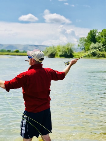 A person wearing a red shirt, black shorts, and a cap is fly fishing in a shallow river. The water is clear, and the person is holding a fishing rod with a line extended. The background features trees and a mountain range under a partly cloudy sky.