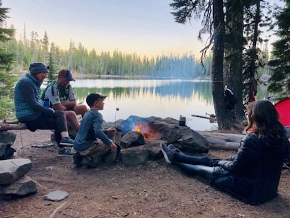 A serene lakeside scene with four people sitting around a campfire. The group is gathered close to the fire, roasting marshmallows, surrounded by tall trees and a calm body of water. The sky is clear, indicating it's either early morning or late evening.