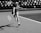 A female tennis player in motion, reaching out with her racket towards a tennis ball. She is dressed in athletic tennis attire and is captured mid-stride on a tennis court with visible lines. The background shows a blurred audience seated in rows of chairs.