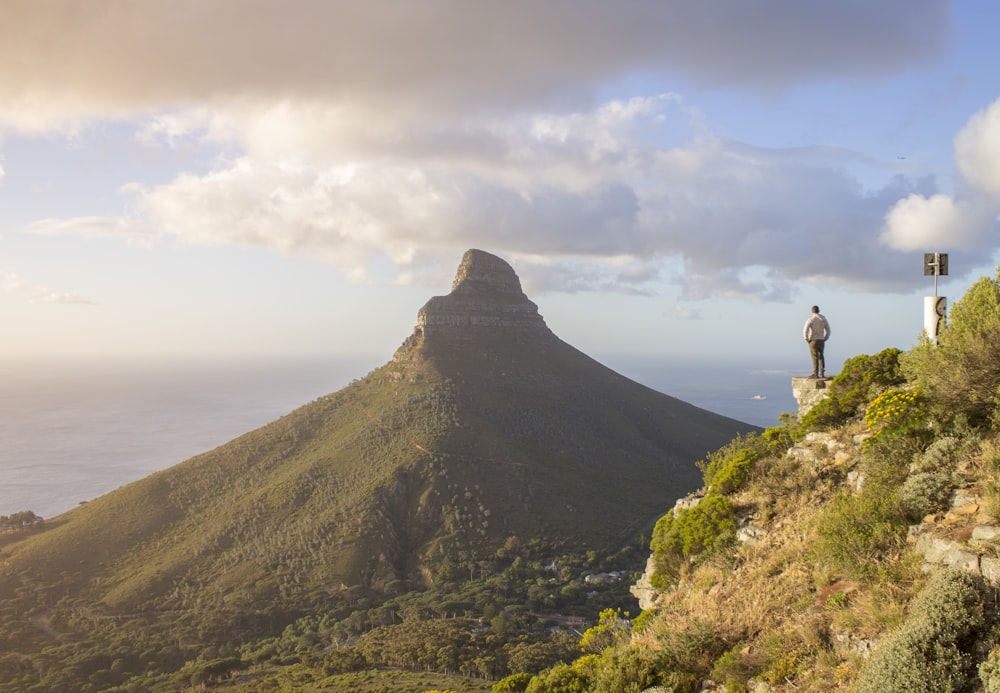 man standing on cliff facing green mountain