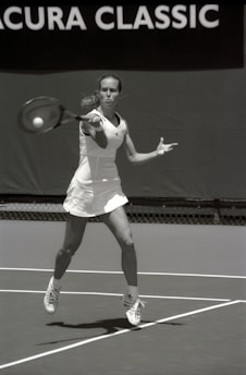 A tennis player wearing a white outfit is captured mid-action on a tennis court, preparing to hit a ball with a forehand stroke. The background shows a banner with the text 'Acura Classic.' The image is in black and white, highlighting the player's focus and motion.