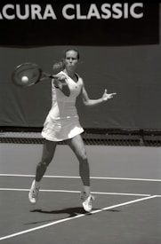 A tennis player wearing a white outfit is captured mid-action on a tennis court, preparing to hit a ball with a forehand stroke. The background shows a banner with the text 'Acura Classic.' The image is in black and white, highlighting the player's focus and motion.