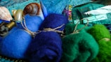 A collection of colorful wool bundles tied with twine, placed alongside seashells and a mechanical spool. The scene is set against a backdrop of textured fabrics and glass bottles, giving an artsy, eclectic feel.