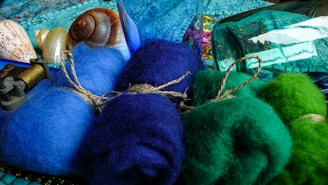 A collection of colorful wool bundles tied with twine, placed alongside seashells and a mechanical spool. The scene is set against a backdrop of textured fabrics and glass bottles, giving an artsy, eclectic feel.