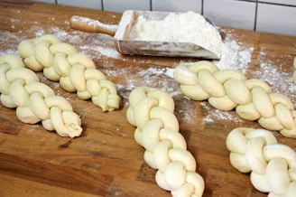 Several pieces of braided dough are placed on a wooden countertop dusted with flour. A metal scoop containing more flour is nearby, suggesting preparation for baking. The dough pieces appear smooth and evenly plaited.