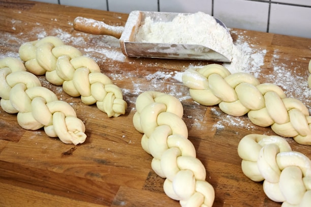 Several pieces of braided dough are placed on a wooden countertop dusted with flour. A metal scoop containing more flour is nearby, suggesting preparation for baking. The dough pieces appear smooth and evenly plaited.