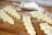 Several pieces of braided dough are placed on a wooden countertop dusted with flour. A metal scoop containing more flour is nearby, suggesting preparation for baking. The dough pieces appear smooth and evenly plaited.