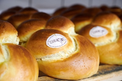 Several freshly baked round bread rolls with a golden brown crust are arranged closely together. Each roll has a small, white label displaying text in the center. The texture of the bread appears soft and fluffy.