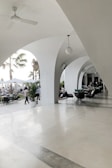 An elegant patio area with white arches creates a sophisticated outdoor setting. Several tables and chairs are arranged for dining, and people are seen both inside the covered area and outside. Palm trees and potted plants contribute to a tropical ambiance. The lighting is bright and natural, suggesting a daytime setting.