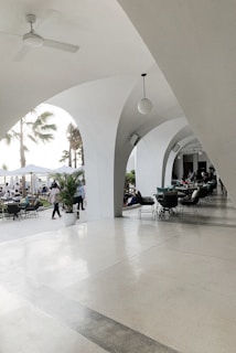 An elegant patio area with white arches creates a sophisticated outdoor setting. Several tables and chairs are arranged for dining, and people are seen both inside the covered area and outside. Palm trees and potted plants contribute to a tropical ambiance. The lighting is bright and natural, suggesting a daytime setting.