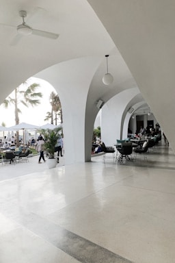 An elegant patio area with white arches creates a sophisticated outdoor setting. Several tables and chairs are arranged for dining, and people are seen both inside the covered area and outside. Palm trees and potted plants contribute to a tropical ambiance. The lighting is bright and natural, suggesting a daytime setting.