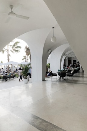 An elegant patio area with white arches creates a sophisticated outdoor setting. Several tables and chairs are arranged for dining, and people are seen both inside the covered area and outside. Palm trees and potted plants contribute to a tropical ambiance. The lighting is bright and natural, suggesting a daytime setting.