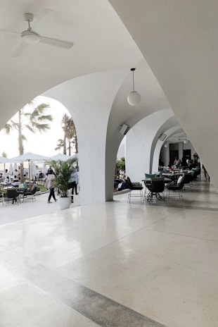 An elegant patio area with white arches creates a sophisticated outdoor setting. Several tables and chairs are arranged for dining, and people are seen both inside the covered area and outside. Palm trees and potted plants contribute to a tropical ambiance. The lighting is bright and natural, suggesting a daytime setting.