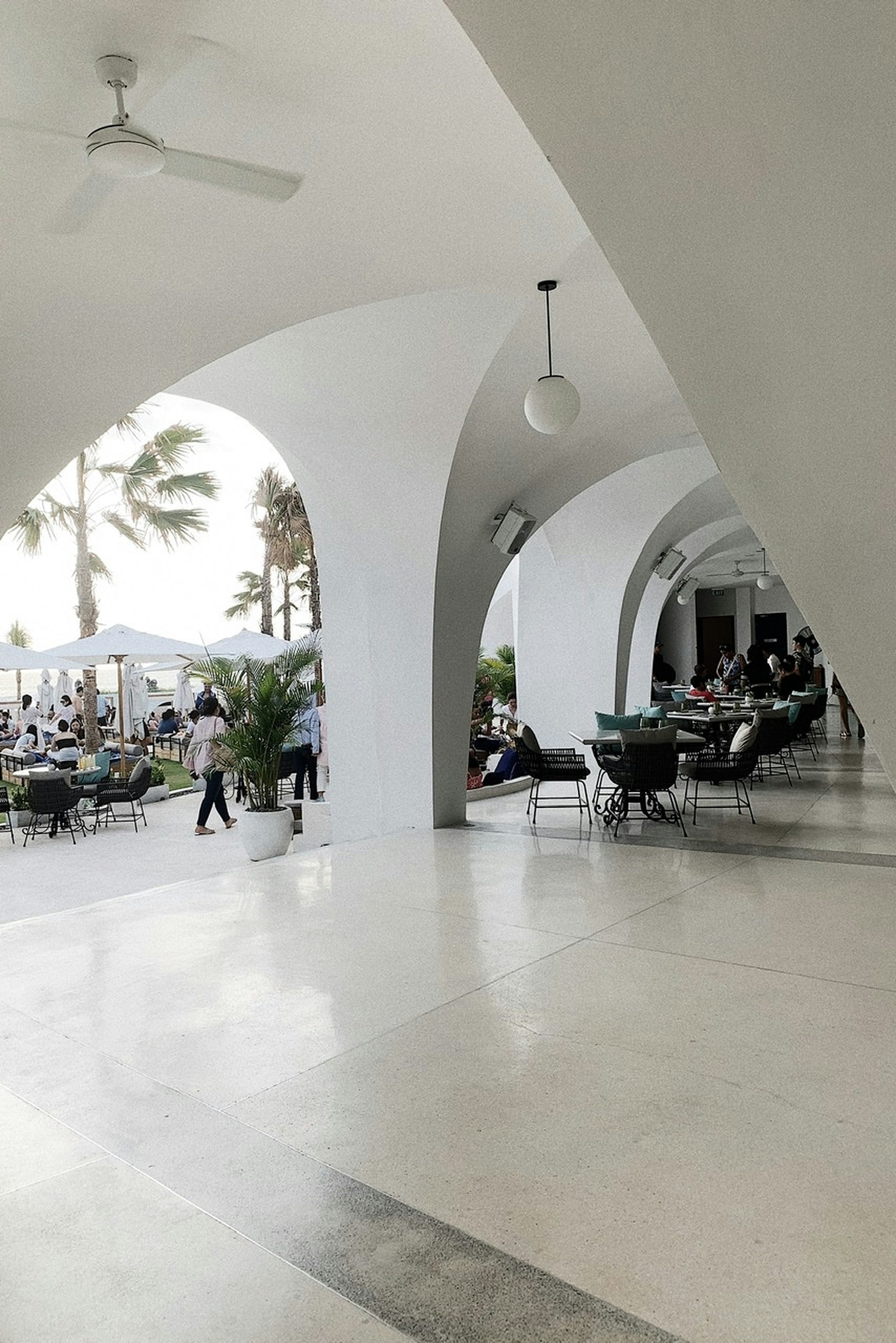 An elegant patio area with white arches creates a sophisticated outdoor setting. Several tables and chairs are arranged for dining, and people are seen both inside the covered area and outside. Palm trees and potted plants contribute to a tropical ambiance. The lighting is bright and natural, suggesting a daytime setting.
