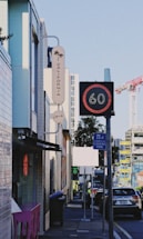 An urban street scene featuring contemporary buildings, a 'California' sign with a palm tree graphic, and a digital traffic sign displaying the number 60. The street includes parked cars, a commercial loading zone sign, and construction cranes visible in the background.