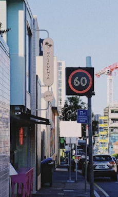 An urban street scene featuring contemporary buildings, a 'California' sign with a palm tree graphic, and a digital traffic sign displaying the number 60. The street includes parked cars, a commercial loading zone sign, and construction cranes visible in the background.