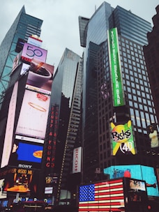 Skyscrapers dominate the urban skyline, adorned with vibrant and large electronic billboards displaying advertisements and brand names. An American flag is prominently displayed in lights.