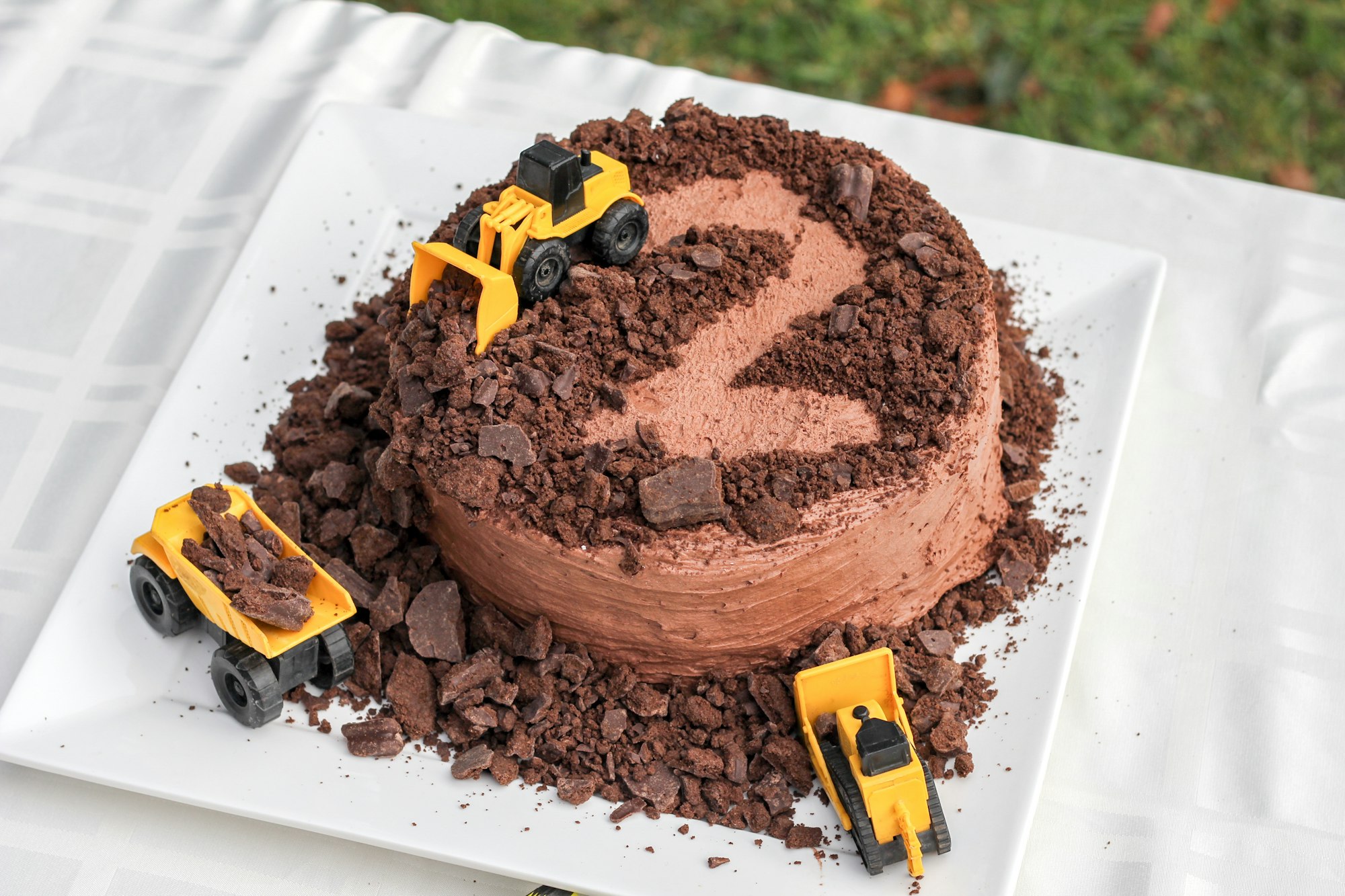 a cake covered in chocolate chunks with the number two scraped out, with three toy construction trucks