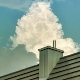 A large, fluffy cloud rises majestically in the sky, framed by a clear, blue background. In the foreground, a modern metal roof with a chimney adds an industrial contrast against the natural softness of the cloud.