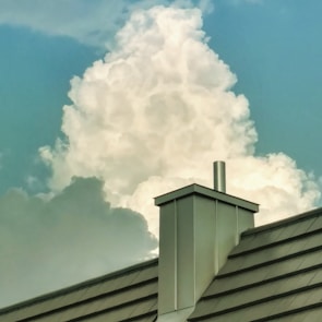 A large, fluffy cloud rises majestically in the sky, framed by a clear, blue background. In the foreground, a modern metal roof with a chimney adds an industrial contrast against the natural softness of the cloud.
