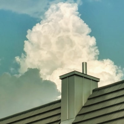 A large, fluffy cloud rises majestically in the sky, framed by a clear, blue background. In the foreground, a modern metal roof with a chimney adds an industrial contrast against the natural softness of the cloud.