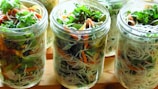Glass jars are filled with vibrant mixed salads composed of noodles, leafy greens, shredded carrots, and various vegetables. The arrangement is visually appealing with layers of colors and textures, showcasing a fresh and healthy meal option.