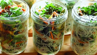 Glass jars are filled with vibrant mixed salads composed of noodles, leafy greens, shredded carrots, and various vegetables. The arrangement is visually appealing with layers of colors and textures, showcasing a fresh and healthy meal option.