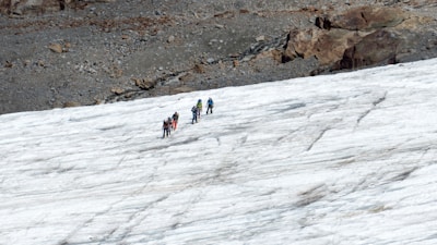 A group of people is trekking across a snowy landscape. They are equipped with climbing gear, likely indicating a challenging hike or mountaineering activity. The terrain is a mixture of ice and rock, with a rugged area visible in the background.