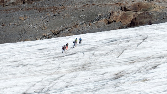 A group of people is trekking across a snowy landscape. They are equipped with climbing gear, likely indicating a challenging hike or mountaineering activity. The terrain is a mixture of ice and rock, with a rugged area visible in the background.