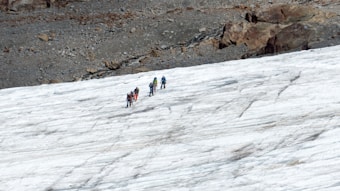 A group of people is trekking across a snowy landscape. They are equipped with climbing gear, likely indicating a challenging hike or mountaineering activity. The terrain is a mixture of ice and rock, with a rugged area visible in the background.