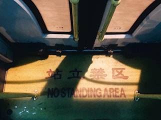 The image depicts the floor of a bus or train with a clear warning sign that reads 'NO STANDING AREA' along with similar text in Chinese characters. The floor is a muted yellow color with visible shadows. Two vertical poles, likely for support, are visible on the left and right sides.