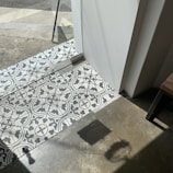 Patterned black and white tiles transition to a concrete floor near a glass door. Sunlight casts sharp shadows on the floor, with part of a table visible in the corner.