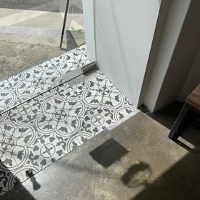 Patterned black and white tiles transition to a concrete floor near a glass door. Sunlight casts sharp shadows on the floor, with part of a table visible in the corner.