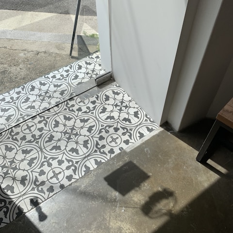 Patterned black and white tiles transition to a concrete floor near a glass door. Sunlight casts sharp shadows on the floor, with part of a table visible in the corner.