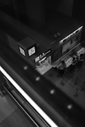 A monochrome image features a currency exchange booth captured from above. The booth displays signs with various symbols, including currency notations, and is attended by several people in uniform standing behind a counter. The scene is framed by a large staircase railing in the foreground, creating a dynamic diagonal across the photo. The setting appears to be inside a building, possibly at an airport or shopping mall.