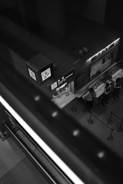A monochrome image features a currency exchange booth captured from above. The booth displays signs with various symbols, including currency notations, and is attended by several people in uniform standing behind a counter. The scene is framed by a large staircase railing in the foreground, creating a dynamic diagonal across the photo. The setting appears to be inside a building, possibly at an airport or shopping mall.