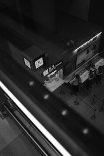 A monochrome image features a currency exchange booth captured from above. The booth displays signs with various symbols, including currency notations, and is attended by several people in uniform standing behind a counter. The scene is framed by a large staircase railing in the foreground, creating a dynamic diagonal across the photo. The setting appears to be inside a building, possibly at an airport or shopping mall.