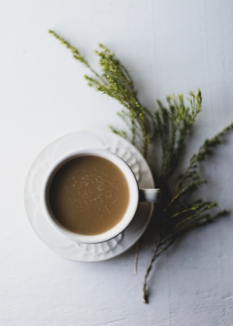 A cup of coffee sits on a light-colored tabletop, accompanied by a white saucer. Green, fern-like sprigs are arranged next to the saucer, providing a natural accent.