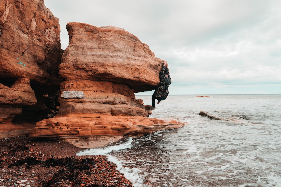 man climbing on brown rock beside beach